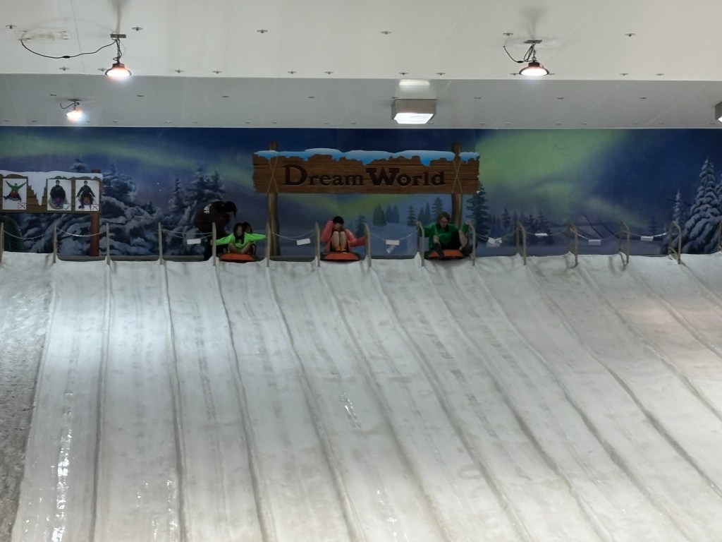 Two young girls in winter jackets sitting at the top of a colourful slide inside the Snow Dome at Dream World Bangkok, Thailand, surrounded by artificial snow and icy scenery.