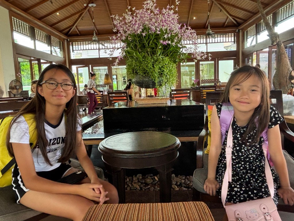 Two smiling girls seated in the spacious, warmly lit lobby of InterContinental Pattaya Resort in Thailand, with plush seating and tropical décor in the background.